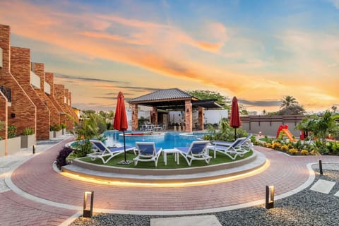 A pool area with lounge chairs, red umbrellas, and lush greenery. A shaded structure with seating, a colorful slide, and a warm sunset sky create a relaxing ambiance.