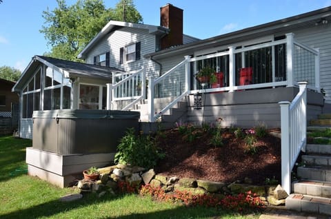 This inviting lakefront home features a raised deck with white railings, cozy seating, and potted plants. Below, a landscaped garden with colorful flowers adds charm, while a hot tub and screened sunroom offer relaxation and comfort.