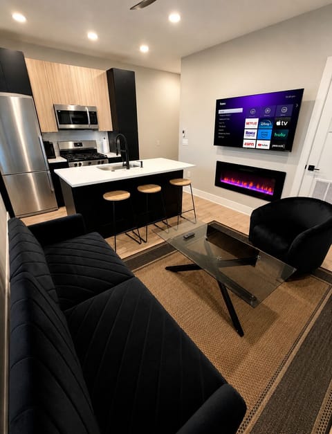 Modern open-concept living space with futon sofa bed, Roku TV, electric fireplace, and fully equipped kitchen with island seating—perfect for relaxing or extra guests.