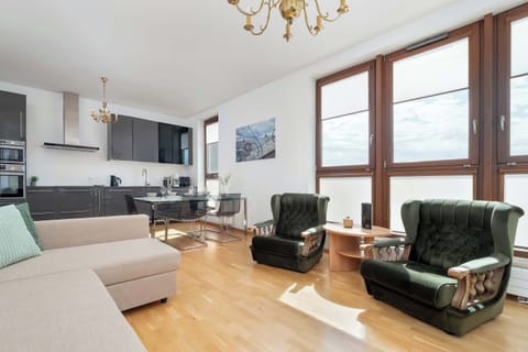 A living room with a cozy sofa, armchairs, and TV area. The wooden floor and light walls create a warm, inviting atmosphere.