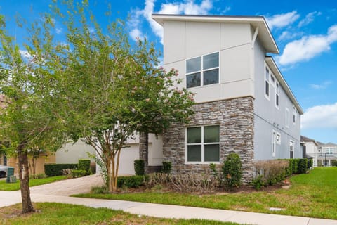 Two-story home exterior features a stone facade, light gray siding, and a brick-paved driveway with surrounding greenery.
