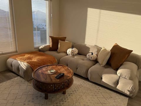 Escape to our cozy retreat. Sink into the cloud couch with a soft blanket and pillow. Natural light from two windows fills the room, highlighting the plush carpet and modern center table. Your perfect getaway awaits.