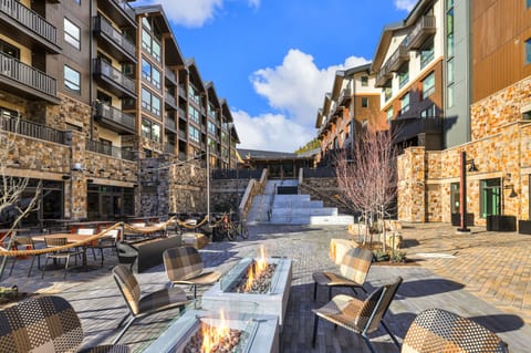 Seating area with fire pits, surrounded by stone buildings and trees.