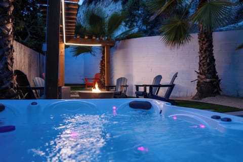 Twilight hot tub with LED jets glowing beside palm trees, fire pit, and Adirondack seating — the ultimate wind-down combo