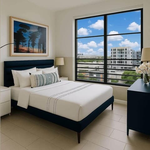 Bedroom 1 of 2 features comfy queen-size bed with luxury linens, floor to ceiling windows offering city views and natural light and also full black out shades. Includes dresser for storage, closet with hangers, and a modern curved LED lamp.