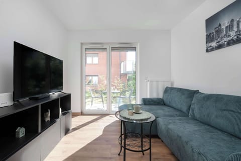 A cozy living room with a green sofa, TV on a stand, and large window letting in natural light. The wooden floor adds warmth to the modern decor.