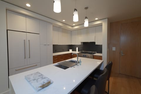 Your sleek modern kitchen awaits with pristine white quartz countertops and warm wood accents. The spacious island invites you to prep meals while pendant lights cast a gentle glow overhead.