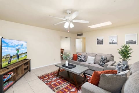 Cozy living area with a large sectional facing the TV, perfect for relaxed movie nights or catching up after a day out. A colorful patterned rug, coffee table centerpiece, and leafy plant add warmth while the ceiling fan keeps the space breezy.