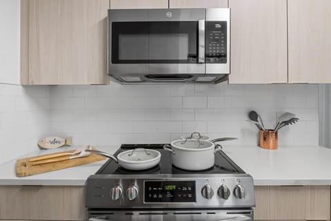 Modern kitchen with stainless steel appliances, white subway tile backsplash, and light wood cabinets. Features a sleek stove, microwave, cookware, and utensils for a clean, stylish cooking space.