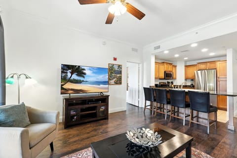 Bright and modern open-concept living space featuring a cozy seating area, large flat-screen TV, and a fully equipped kitchen with a breakfast bar. Hardwood floors and contemporary lighting create a warm and inviting atmosphere.