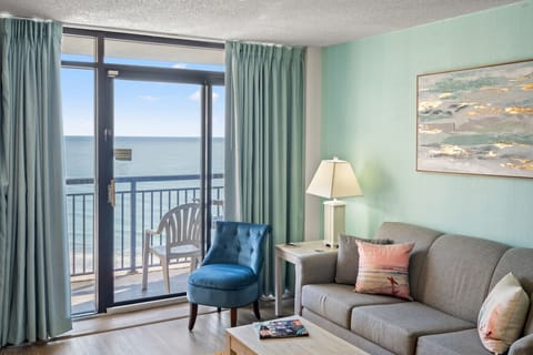 Expansive oceanfront living room with panoramic Atlantic views, plush queen sleeper sofa, and Smart TV. Open kitchen makes meals easy, while sliding doors lead to a private balcony overlooking the beach and coastline.