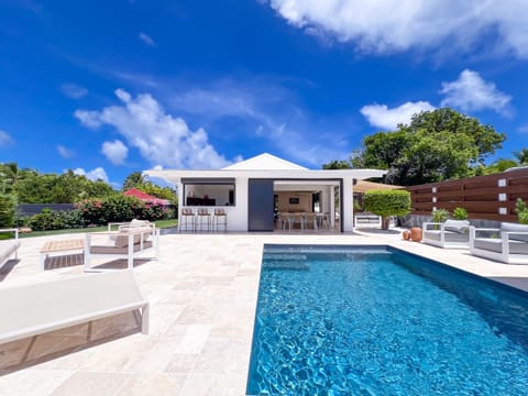 Large pool with sun loungers and outdoor lounge, facing a modern house under the tropical blue sky.