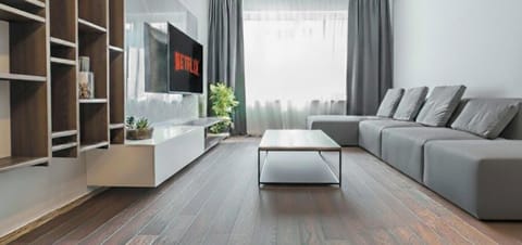 Modern living room with a large gray sectional, coffee table on a light rug, wall-mounted TV, shelves, and a plant. Bright space with natural light from large windows and dark curtains.