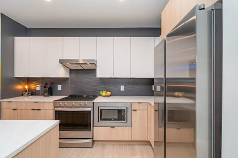 Full kitchen with stainless steel appliances, pristine white countertops, light wood and white cabinetry, and a spacious island with breakfast bar seating for four.