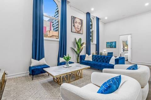 Bright and elegant living room with plush blue velvet sofas, marble coffee table, tall arched windows, and city views. A stylish space designed for comfort, natural light, and relaxed conversations.