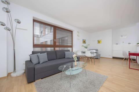 A bright living room with a gray sofa, TV, and large window letting in natural light. The space is decorated in a modern style with a light rug and dining table in the background.