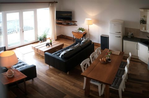 View from the stairs towards the living and kitchen area; dining table with six chairs, fridge/freezer, sofas, coffee table, lamps and TV