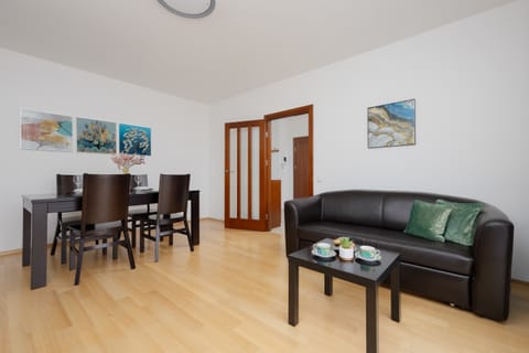 A spacious living room with a black sofa, dining table, and wooden flooring. Colorful paintings add personality to the bright space.