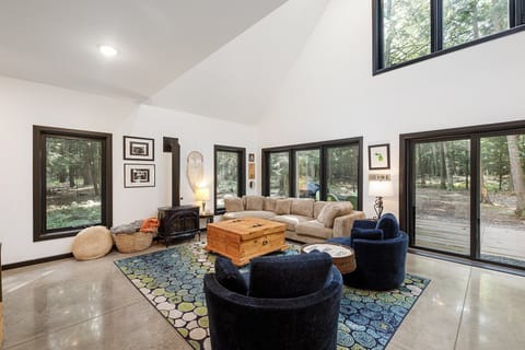 Sun-filled living room w/ soaring ceilings, cozy seating, & walls of windows framing peaceful woodland views.