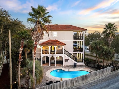 Three-story house with red tile roof, arched balconies, and a kidney-shaped pool surrounded by palm trees.