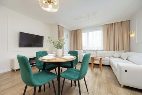 A modern dining and living space with a wooden table, green upholstered chairs, and a white sofa. Large windows brighten the room with natural light.