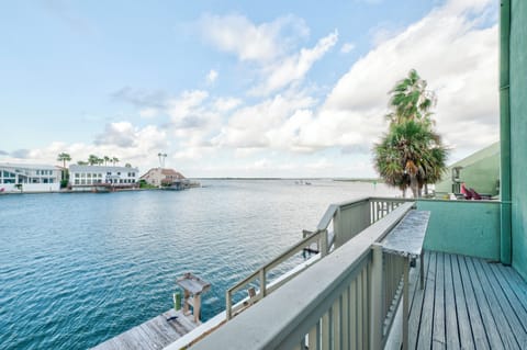 Waterfront balcony overlooking tranquil bay waters with palm trees and coastal homes dotting the shoreline.