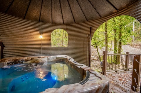 A luxurious hot tub enclosed in a rustic metal gazebo with ambient lighting, offering a warm and inviting spa experience. Relax in the bubbling water while enjoying the surrounding forest view