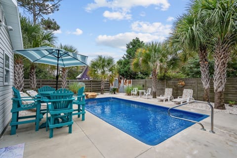 Sunlit Backyard Oasis w/ Sparkling Blue Pool, Palm Trees, Cozy Patio Setup, Turquoise Chairs and Shaded Table with a Striped Umbrella