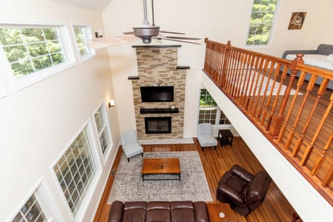 You'll love gathering in this stunning two-story living room, where the soaring stone fireplace creates a cozy focal point. Sink into plush leather seating under vaulted ceilings, with sunlight streaming through tall windows revealing lush treetop views.