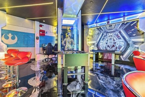 Enter a lively game room with bold blue and red accents, sleek bar stools, and a chic foosball table. The glossy black floor reflects vibrant lighting, setting the perfect atmosphere for fun and relaxation.