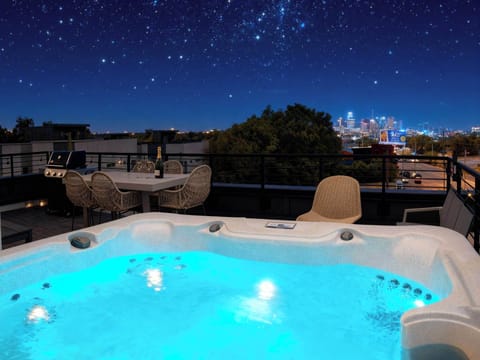 Blue-glowing hot tub on the rooftop with the city skyline behind you, a dining table steps away and a grill ready to fire up. Book your stay with Misfit Homes to claim both houses for your crew