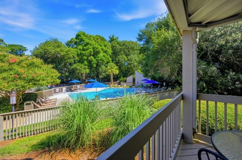 Verdant trees frame a sparkling pool area, seen directly from the private comfort of your shaded balcony. Cobalt umbrellas pop against the bright afternoon sky, offering a cheerful invitation to slow down and embrace the fresh air.