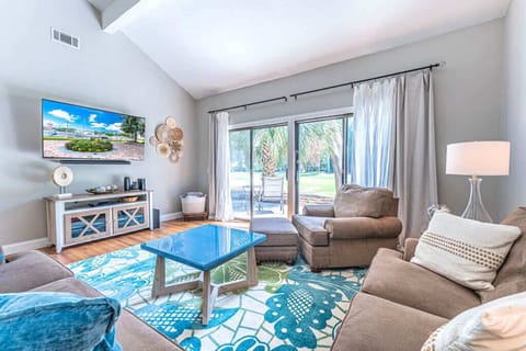 Bright, airy living room with vaulted ceilings, hardwood floors, and a smart TV. Coastal-inspired décor, cozy seating, and sliding glass doors opening to a private patio with golf course views.