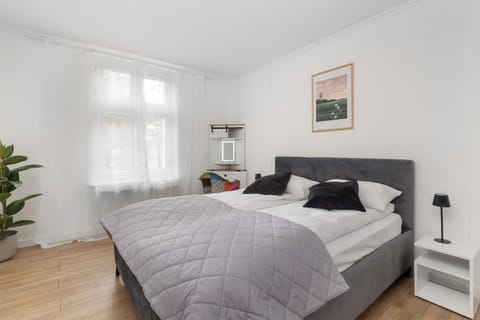 A bedroom featuring a double bed with a grey bedspread, positioned near a large window. Light-colored walls enhance the sense of space
