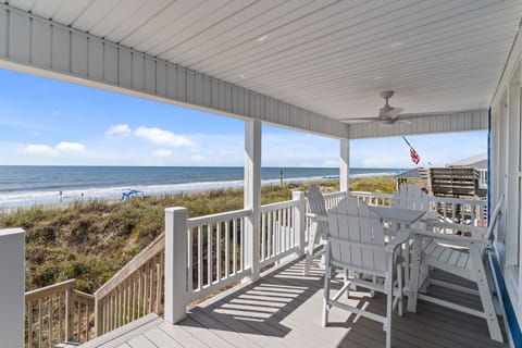 Covered porch overlooking ocean with white railing, Adirondack chairs, dining table, ceiling fan, stairway to dunes, grassy dunes, sandy beach, rolling waves, and neighboring deck with flag.