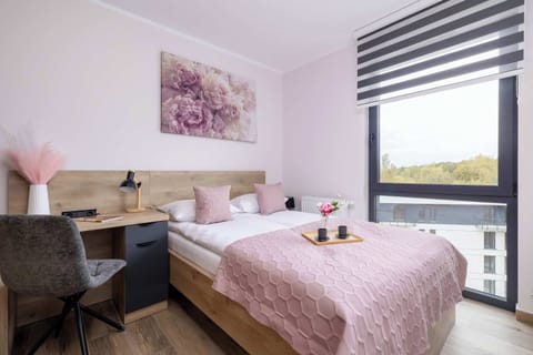 A bedroom features a bed with pink bedding, nightstand, and a large window providing ample natural light.