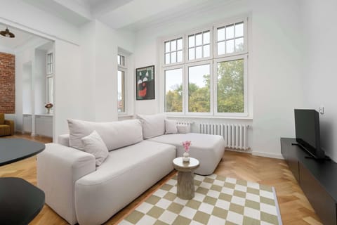 A stylish living room with white walls, wooden flooring, and a checkered rug. Large windows allow natural light to fill the cozy seating area.