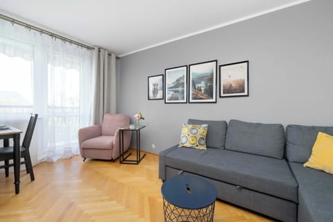 A cozy living room with a gray sectional sofa, framed artwork, and a dining table near the window. Natural light enhances the elegant simplicity.