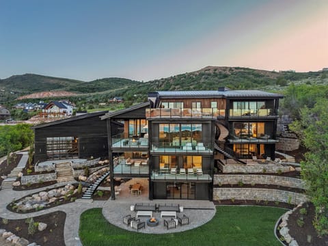 The Halai, an architectural masterpiece set against rolling green hills, featuring expansive terraces, an outdoor fire pit gathering area, and contemporary design that harmonizes with the natural mountain landscape and complements Bear Lake.