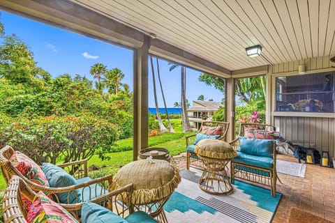 Covered outdoor lanai with cozy wicker seating, vibrant cushions, and a tropical garden backdrop overlooking the sparkling blue ocean, perfect for enjoying morning coffee or sunset cocktails