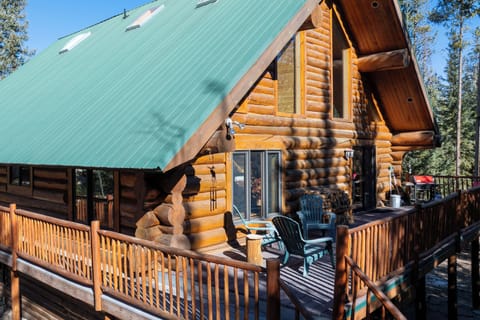 This rustic log cabin features a distinctive green metal roof and wraparound deck, nestled among towering pine trees in a peaceful forest setting.