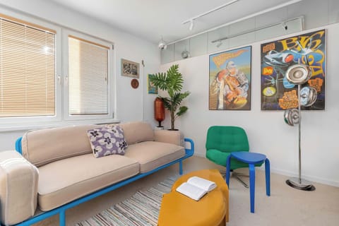 A bright living room with a beige sofa, colorful wall art, and modern decor. The room has a cozy and artistic atmosphere.