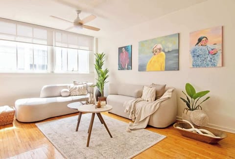 A sunlit living room where modern design meets artistic charm. The soft neutral sofas, warm wooden floors, and vibrant paintings create a welcoming space to relax, unwind, and feel inspired.