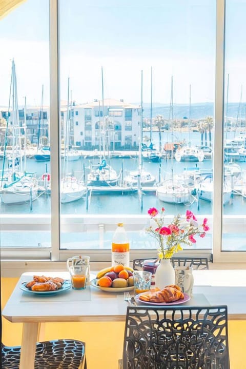 Petit-déjeuner avec vue sur le port de Leucate – table dressée avec fruits frais, jus d`orange et fleurs roses encadrées par la vue panoramique sur les voiliers. Un réveil de rêve au bord de l`eau.