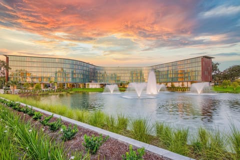 Hotel ELEO’s sleek architecture reflects the vibrant sunset over the serene pond, complete with elegant fountains that enhance the inviting ambiance.