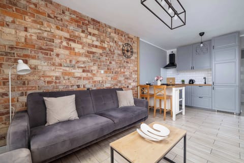 A cozy living room with a gray sofa and a small coffee table, featuring exposed brick walls for a warm atmosphere. The kitchenette is visible in the background.
