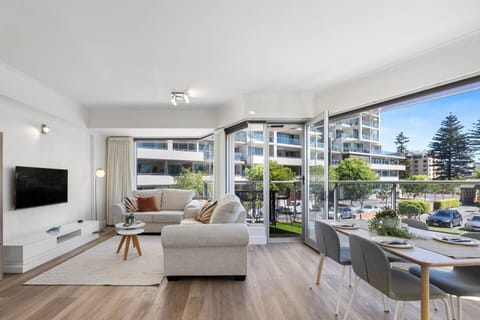 Sunlight pours through wide windows into the open-plan living area, where soft neutral tones, a wall-mounted TV, and a plush sofa create a calm space to unwind.
