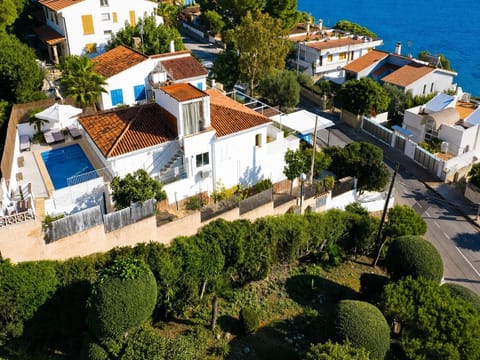 An aerial view showcases a multi-level white villa with traditional terracotta tile roofs, featuring a private swimming pool, sun loungers, and an elevated terrace just a short distance from the blue sea.