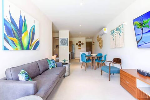 Bright and modern living room with vibrant décor, comfy seating, smart TV, Bose speaker, air conditioning, and a ceiling fan. The open layout connects to the dining area and kitchen, creating a relaxing space after a day at the beach.