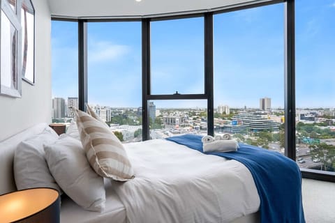 A bright bedroom with curved floor-to-ceiling windows wrapping around the bed, offering sweeping views across Melbourne`s inner suburbs under a clear blue sky. White linen, striped cushions, and a navy throw complete the relaxed, airy feel.